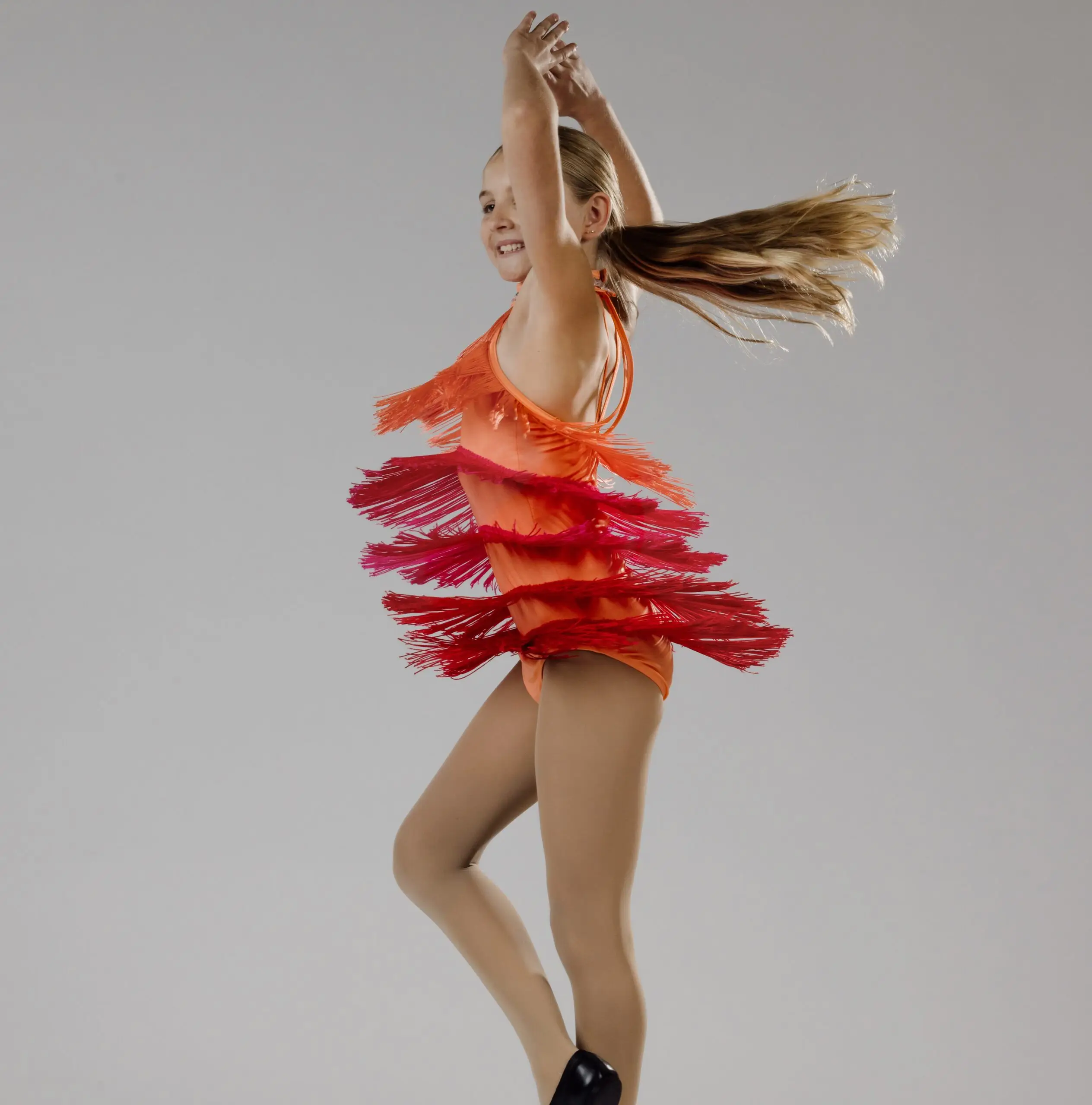 Quince and Mulberry Studios_BaysideDance scaled Young jazz dancer in an orange and red fringe costume spinning with arms raised, smiling and full of energy on a studio background.